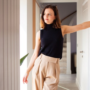 May include: A woman wearing a black sleeveless turtleneck top and beige wide-leg pants. She is standing in a hallway with a white door behind her.