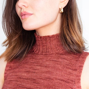 May include: A close-up of a woman wearing a rust-colored sleeveless turtleneck sweater. The sweater has a high neckline and a textured knit.
