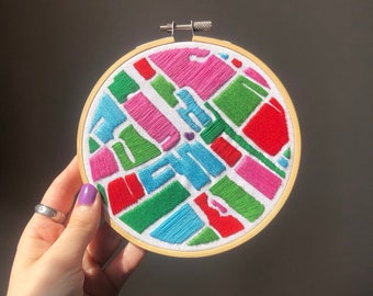 May include: A colorful embroidery hoop featuring an abstract design with the word "Austin" stitched in blue. The design includes pink, green, and red geometric shapes on a white background, set within a wooden hoop.