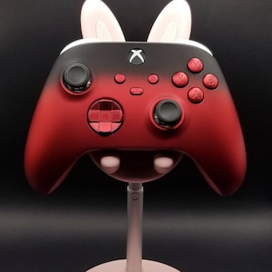 May include: A red and black Xbox controller with bunny ears on top. The controller is on a pink stand.