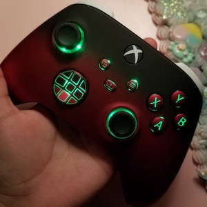 May include: A black and red Xbox controller with green LED lights around the buttons and a textured grip. The controller has a black and white Xbox logo on the front.