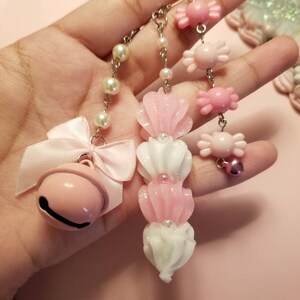 以下が含まれることがあります： ベル、キャンディー、花の形をしたチャームが付いたピンクと白のチャームチェーン2本。