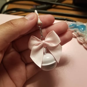 May include: A white bell charm with a pink satin bow. The bell has a silver chain attached to a white plastic clip.