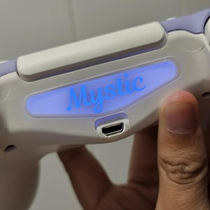 May include: A white and lavender game controller with the word "Mystic" illuminated in blue. The controller has a USB port and is held by a hand.