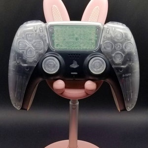 May include: A clear plastic PlayStation 5 controller with pink bunny ears attached to the top. The controller is on a pink stand.