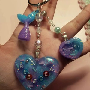 以下が含まれることがあります： 青と紫のマーメイドテールのチャームと、カラフルな文字で「REBECCA」と書かれたハート型のチャーム。チャームは、クリアとパールカラーのビーズが付いたシルバーチェーンに繋がっています。