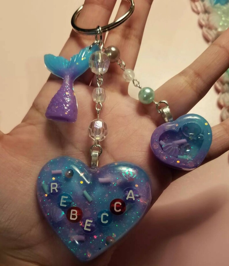以下が含まれることがあります： 青と紫のマーメイドテールのチャームと、カラフルな文字で「REBECCA」と書かれたハート型のチャーム。チャームは、クリアとパールカラーのビーズが付いたシルバーチェーンに繋がっています。