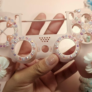 May include: A pink and white custom controller for a video game console, decorated with rhinestones and small charms. The controller has a pink and white design with a floral pattern.