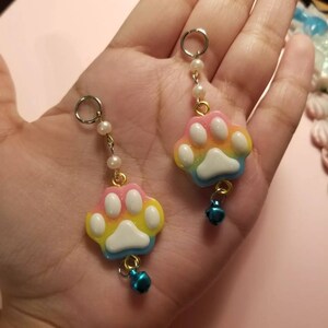 以下が含まれることがあります： 虹色の肉球のモチーフのイヤリング。白いアクセントと青いベルが付いています。ポリマークレイで作られており、銀色のチェーンに白いビーズが通っています。