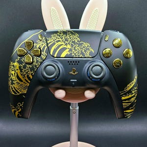 May include: Black and gold PlayStation 5 controller with a wave design and gold buttons. The controller has a bunny ear attachment. The controller is on a pink stand.