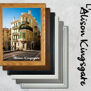 May include: A framed photograph of a yellow building with green shutters and balconies. The building is on a street with a blue sky in the background. The frame is made of wood and has a light brown finish. The photo is signed "Alison Kingsgate".