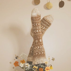 Puede incluir: Un par de calcetines de punto marrón y blanco con un patrón de luna y estrellas. Los calcetines están colgados con las puntas hacia el techo.