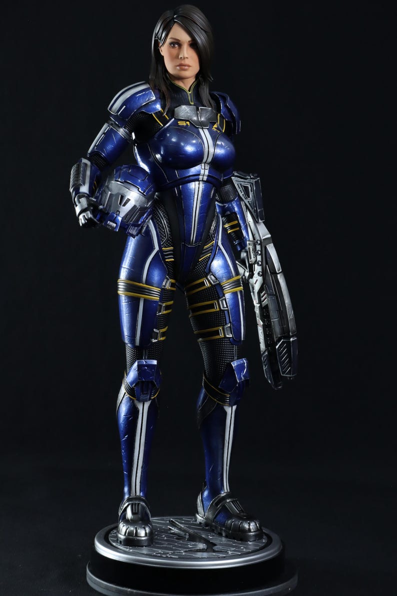 May include: A blue and white action figure of a woman in a futuristic suit, holding a weapon. The figure is standing on a black base.