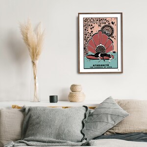 May include: A framed print of a goddess with a crown of flowers, lying on a seashell. The print is in a brown frame and has the text "APHRODITE THE GODDESS OF LOVE" at the bottom.