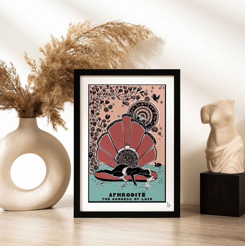 May include: A black framed print featuring a pink and black illustration of a woman reclining on a shell. The text "APHRODITE THE GODDESS OF LOVE" is at the bottom of the print.