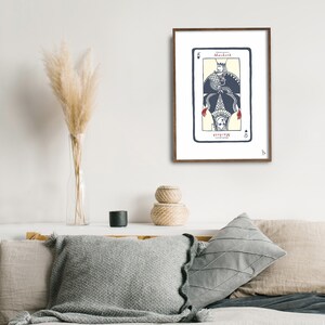 May include: A framed print of a playing card with a black spade suit and the word "Macbeth" printed on it. The card features a stylized illustration of a person wearing a crown and a robe with a bird perched on their head.