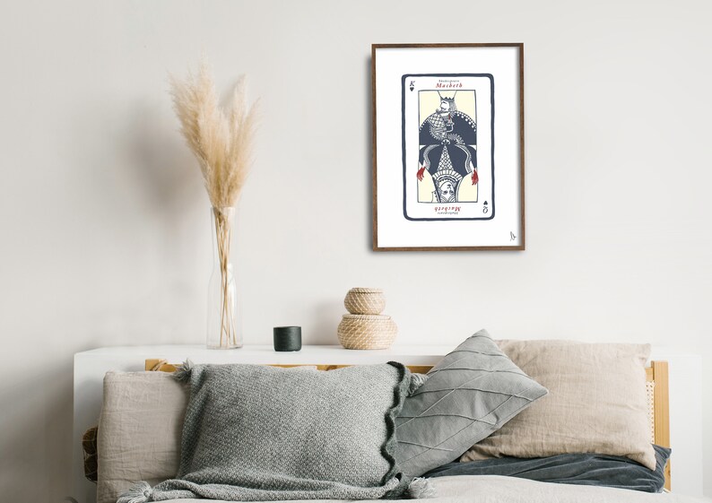 May include: A framed print of a playing card with a black spade suit and the word "Macbeth" printed on it. The card features a stylized illustration of a person wearing a crown and a robe with a bird perched on their head.