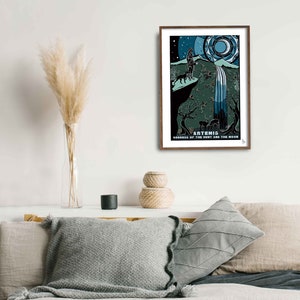 May include: A framed print of a stylized illustration of Artemis, the Greek goddess of the hunt and the moon. The illustration depicts Artemis standing on a cliff with a bow and arrow, a wolf at her side, and a waterfall cascading down the cliff. The background is a dark blue sky with stars and a crescent moon.