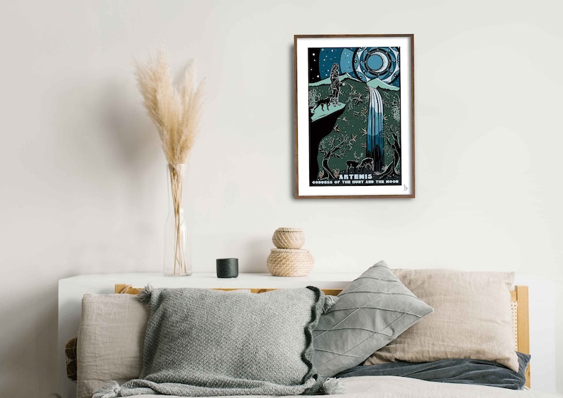 May include: A framed print of a stylized illustration of Artemis, the Greek goddess of the hunt and the moon. The illustration depicts Artemis standing on a cliff with a bow and arrow, a wolf at her side, and a waterfall cascading down the cliff. The background is a dark blue sky with stars and a crescent moon.
