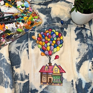 Può includere: Giacca di jeans con un design colorato raffigurante una casa sollevata da palloncini. La giacca ha un effetto sbiancato e tie-dye. È visibile una tavolozza con vernice e pennelli.