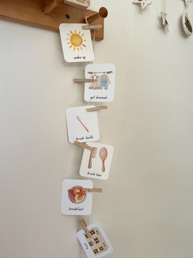 May include: A set of white cards with illustrations and text on them, hung on a wooden wall with clothespins. The cards show a sun, clothes on a clothesline, a toothbrush, a hairbrush, a plate of breakfast food, and a house. The text on the cards reads "wake up", "get dressed", "brush teeth", "brush hair", "breakfast", and "play".