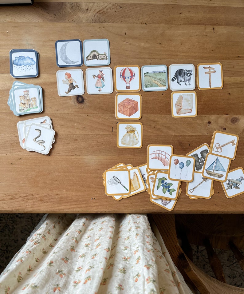 May include: A set of colourful illustrated cards with various images, including a castle, a snake, a hot air balloon, a bridge, a raccoon, a key, a net, a bag, a package, a house, a moon, a cloud with rain, a road, a signpost, and a pair of boots. The cards are laid out on a wooden surface.