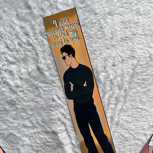 May include: A bookmark with an illustration of a man in sunglasses, black shirt, and trousers. The bookmark has the text "I was always meant to find my way to you" in white font. The background is a gradient of gold and orange.