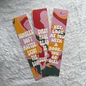 Peut inclure: Trois marque-pages colorés avec des motifs en spirale abstraits. Les marque-pages comportent des citations inspirantes dans une police rétro. Le premier marque-page dit "Les chiffres n'ont pas d'importance, profitez du livre". Le deuxième marque-page dit "Chaque page lue est une suggestion". Le troisième marque-page dit "Hé, regarde-toi avec un livre. Vas-y, fille."