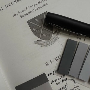 May include: A close-up of a book with the title "The Necessity of Violence" and a crest for the Royal Institute of Translation, University of Oxford. The book is open to a page with the text "An Arcane History of the Oxford Translators' Revolution". A black pen and a gray color chart are also visible.