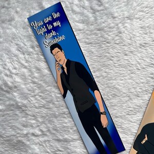 May include: A bookmark with a blue background and the text "You are the light to my dark, Sunshine." The bookmark features an illustration of a person in a black vest and black trousers, holding a phone to their ear.