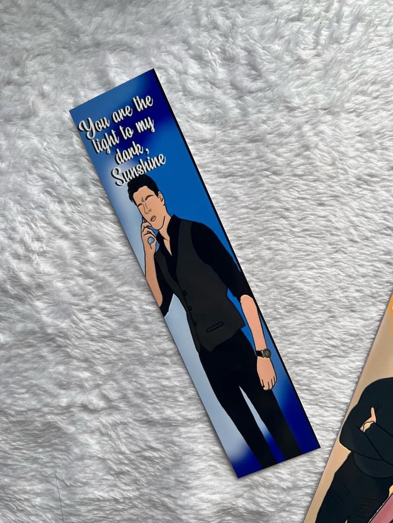 May include: A bookmark with a blue background and the text "You are the light to my dark, Sunshine." The bookmark features an illustration of a person in a black vest and black trousers, holding a phone to their ear.