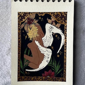 May include: An original painting on a spiral-bound sketchbook. The artwork features a figure with white wings and a flower-like head, set against a black background with gold accents and a decorative border. A red flower is at the bottom.