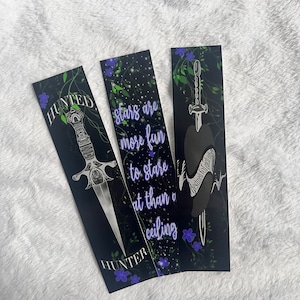 May include: Three black bookmarks with a white floral design and a silver dagger. The bookmarks have the text "HUNTED", "HUNTER", and "stars are more fun to stare at than a ceiling".