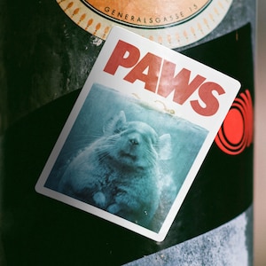 May include: A black metal pole with three stickers. The top sticker is a round, orange and black sticker with the word "Dude" in white. The middle sticker is a white sticker with a picture of a gray animal and the word "PAWS" in red. The bottom sticker is a partially visible pink sticker.