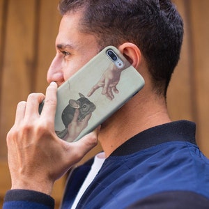 May include: A phone case with a chinchilla illustration, inspired by Michelangelo's The Creation of Adam. The design shows a chinchilla held in a hand, with another hand reaching towards it. The case has a light beige background.