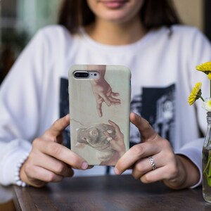 May include: A phone case featuring a Renaissance-style painting of two hands reaching towards each other. The case has a light beige background with the artwork in muted tones of brown, beige, and red. The phone case is held by a person.