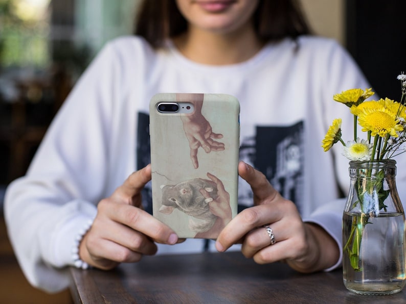 May include: A phone case featuring a Renaissance-style painting of two hands reaching towards each other. The case has a light beige background with the artwork in muted tones of brown, beige, and red. The phone case is held by a person.