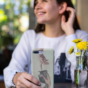 May include: A phone case with a Renaissance-style image of two hands reaching out, with a chinchilla in the middle. The case has a light green background. The phone case is held in front of a blurred background.
