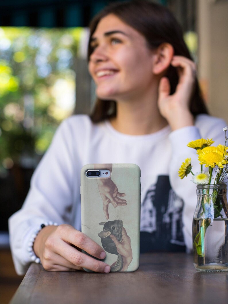 May include: A phone case with a Renaissance-style image of two hands reaching out, with a chinchilla in the middle. The case has a light green background. The phone case is held in front of a blurred background.
