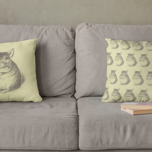 May include: Two decorative throw pillows on a gray sofa. One pillow features a detailed pencil drawing of a chinchilla. The other pillow has a repeating pattern of chinchilla drawings. The pillows are light yellow.