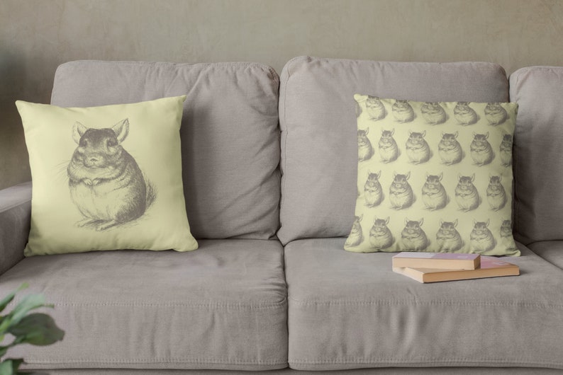 May include: Two decorative throw pillows on a gray sofa. One pillow features a detailed pencil drawing of a chinchilla. The other pillow has a repeating pattern of chinchilla drawings. The pillows are light yellow.