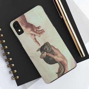 May include: A phone case with a humorous design based on Michelangelo's 'The Creation of Adam,' replacing Adam with a chinchilla. The case has a beige and white background. The phone case is on a black notebook.