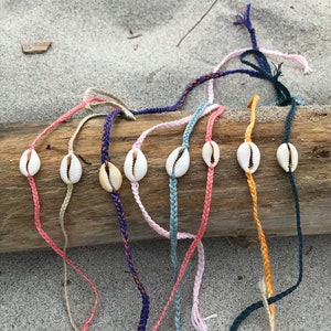 Peut inclure: Plusieurs bracelets faits à la main avec des coquillages cauris et des cordons tressés colorés. Les bracelets sont présentés sur un morceau de bois flotté sur un fond de sable. Chaque bracelet a une couleur unique, comme le rose, le bleu et l'orange.