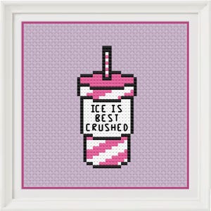 May include: Cross-stitch art in a white frame. The artwork features a pixelated pink and white drink cup with a straw. The text "ICE IS BEST CRUSHED" is displayed on the cup. The background is a light purple fabric.