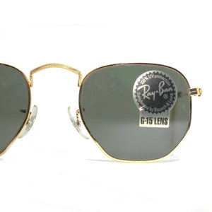 Może przedstawiać: Okulary przeciwsłoneczne w złotej oprawie z zielonymi szkłami. Okulary mają naklejkę na prawym szkle z napisem "100% UV protection Ray-Ban Sunglasses by Bausch & Lomb G-15 LENS".