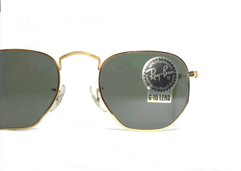Może przedstawiać: Okulary przeciwsłoneczne w złotej oprawie z zielonymi szkłami. Okulary mają naklejkę na prawym szkle z napisem "100% UV protection Ray-Ban Sunglasses by Bausch & Lomb G-15 LENS".