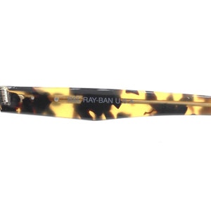 May include: Close-up of the tortoiseshell frame of a pair of Ray-Ban sunglasses. The frame has a gold metal bar and the text "RAY-BAN U.S.A." is printed on the frame.