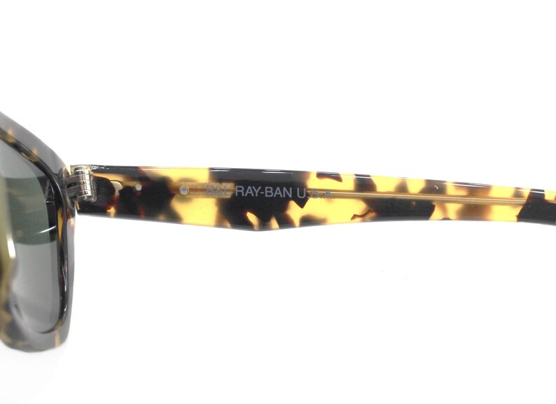 May include: Close-up of the tortoiseshell frame of a pair of Ray-Ban sunglasses. The frame has a gold metal bar and the text "RAY-BAN U.S.A." is printed on the frame.