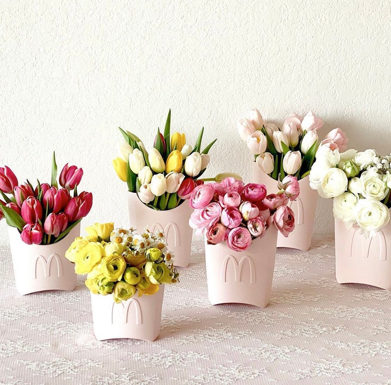 以下が含まれることがあります： ピンクと白のセラミック容器に入ったピンクの花の飾り付け。花には、ピンクのチューリップ、白いチューリップ、黄色のチューリップ、ピンクのバラが含まれています。
