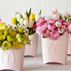 以下が含まれることがあります： 白いMのロゴが付いたピンクの花瓶が3つ。それぞれの花瓶には異なる花がアレンジされています。花はピンク、白、黄色です。